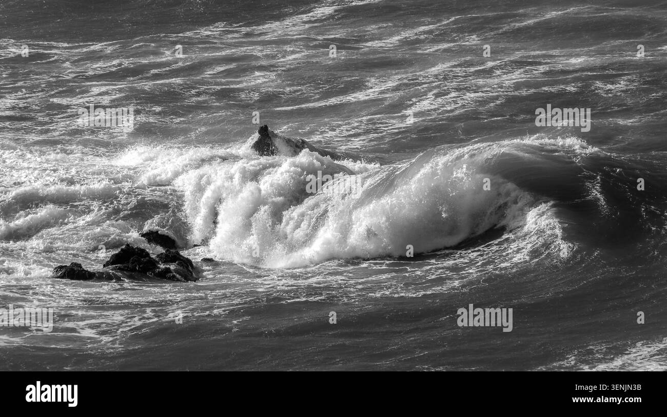 Vague noire et blanche s'écrasant sur les rochers côtiers, créant un paysage marin côtier monochrome spectaculaire. Banque D'Images