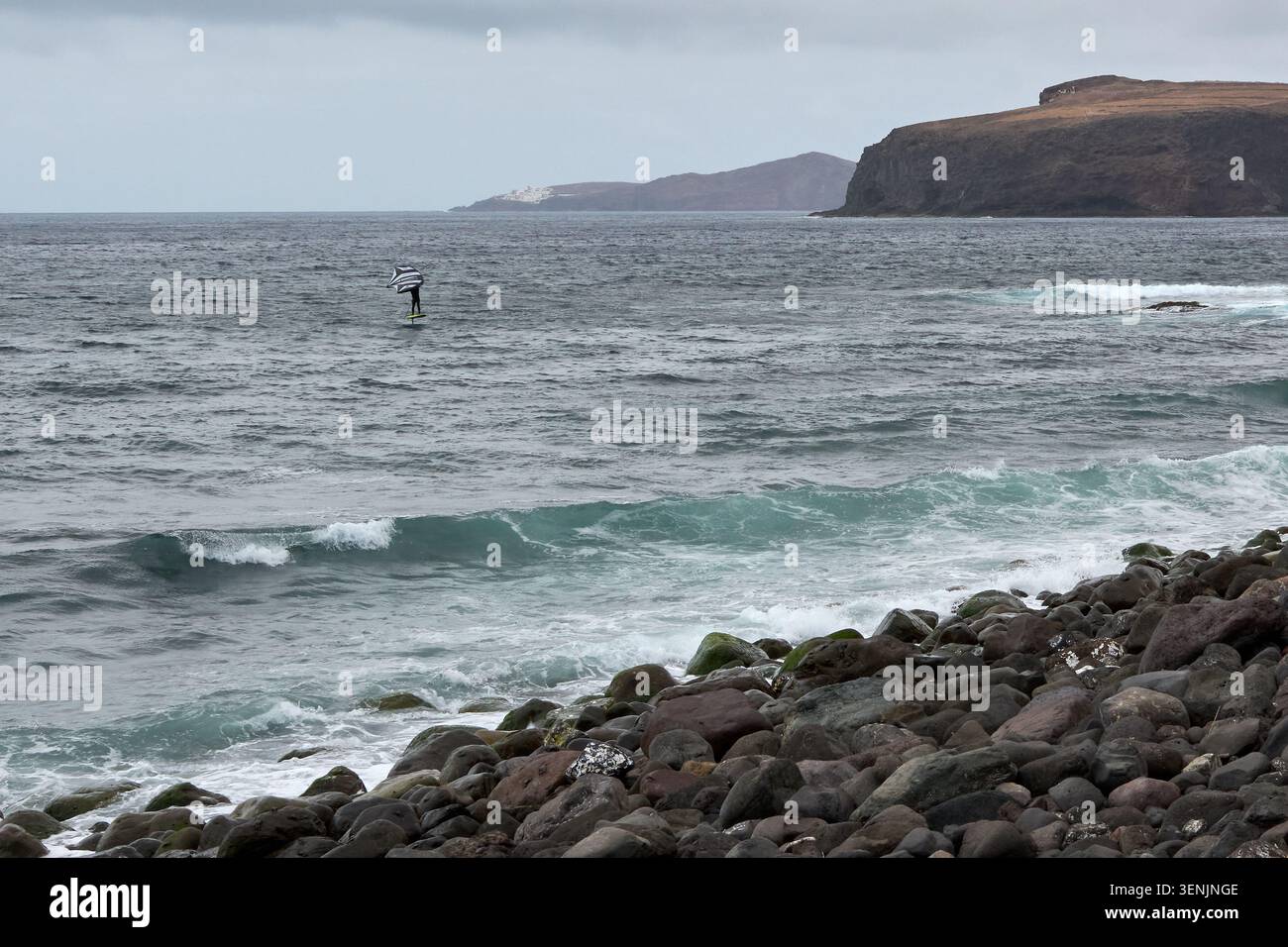 Un athlète pratique le footing d'aile sur l'océan près d'un rivage volcanique rocheux avec des falaises côtières et des collines lointaines sous un ciel couvert. Banque D'Images