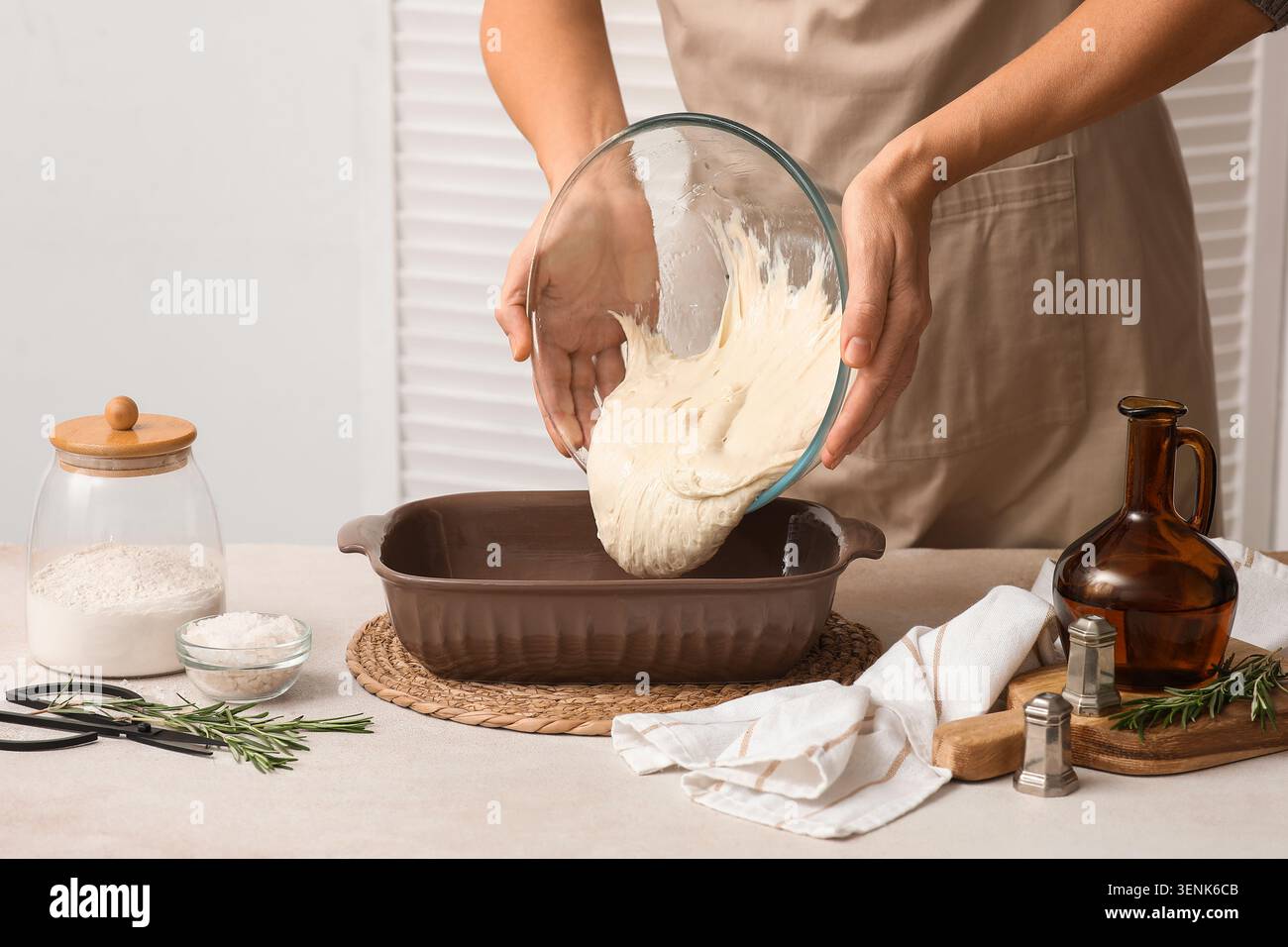 Femme versant la pâte fraîchement préparée du bol en verre dans le plat de cuisson pour le pain ou la pâtisserie maison, divers ingrédients de cuisson et outils sont disposés Banque D'Images