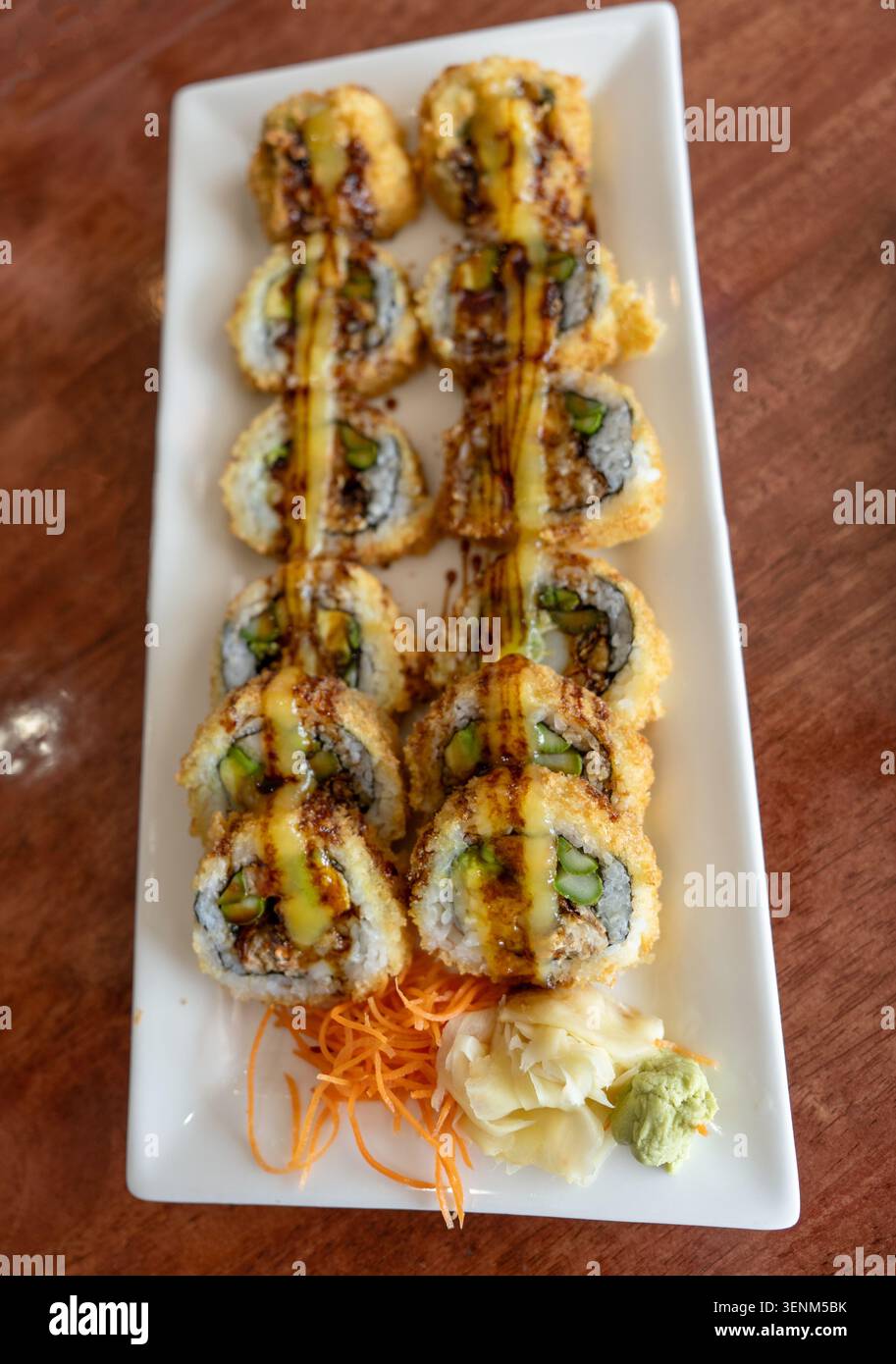 Gros plan de rouleaux de sushi tempura frits croustillants disposés sur une assiette rectangulaire blanche. Les rouleaux sont arrosés de sauce salée et serv Banque D'Images