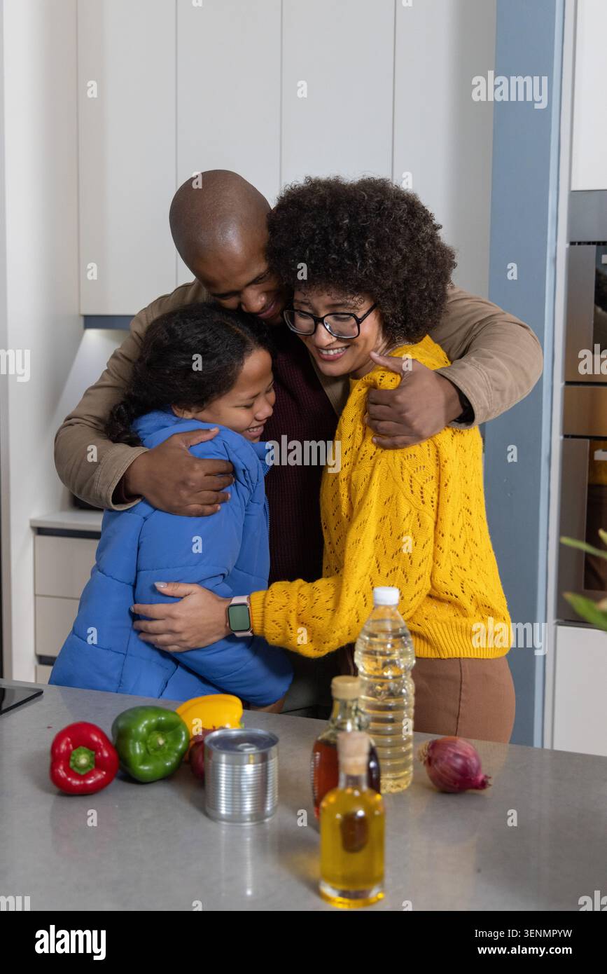 Famille embrassant derrière l'îlot de cuisine avec des poivrons, femme portant un tricot jaune, enfant en soufflet bleu Banque D'Images