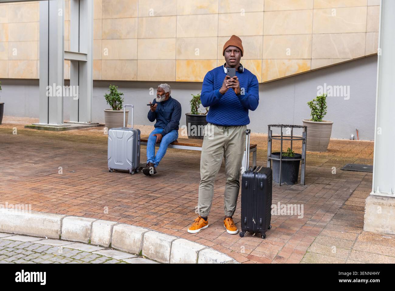 Hommes afro-américains attendant à l'abri à l'extérieur du bâtiment, debout avec le téléphone et des valises roulantes Banque D'Images