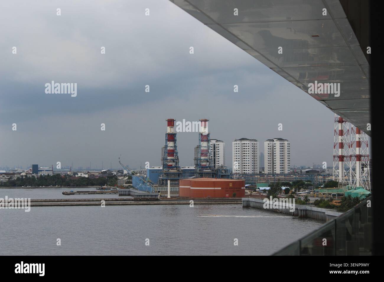 Centrale électrique industrielle avec des cheminées rouges à rayures blanches surplombant un réservoir orange massif à côté des eaux côtières calmes. Banque D'Images
