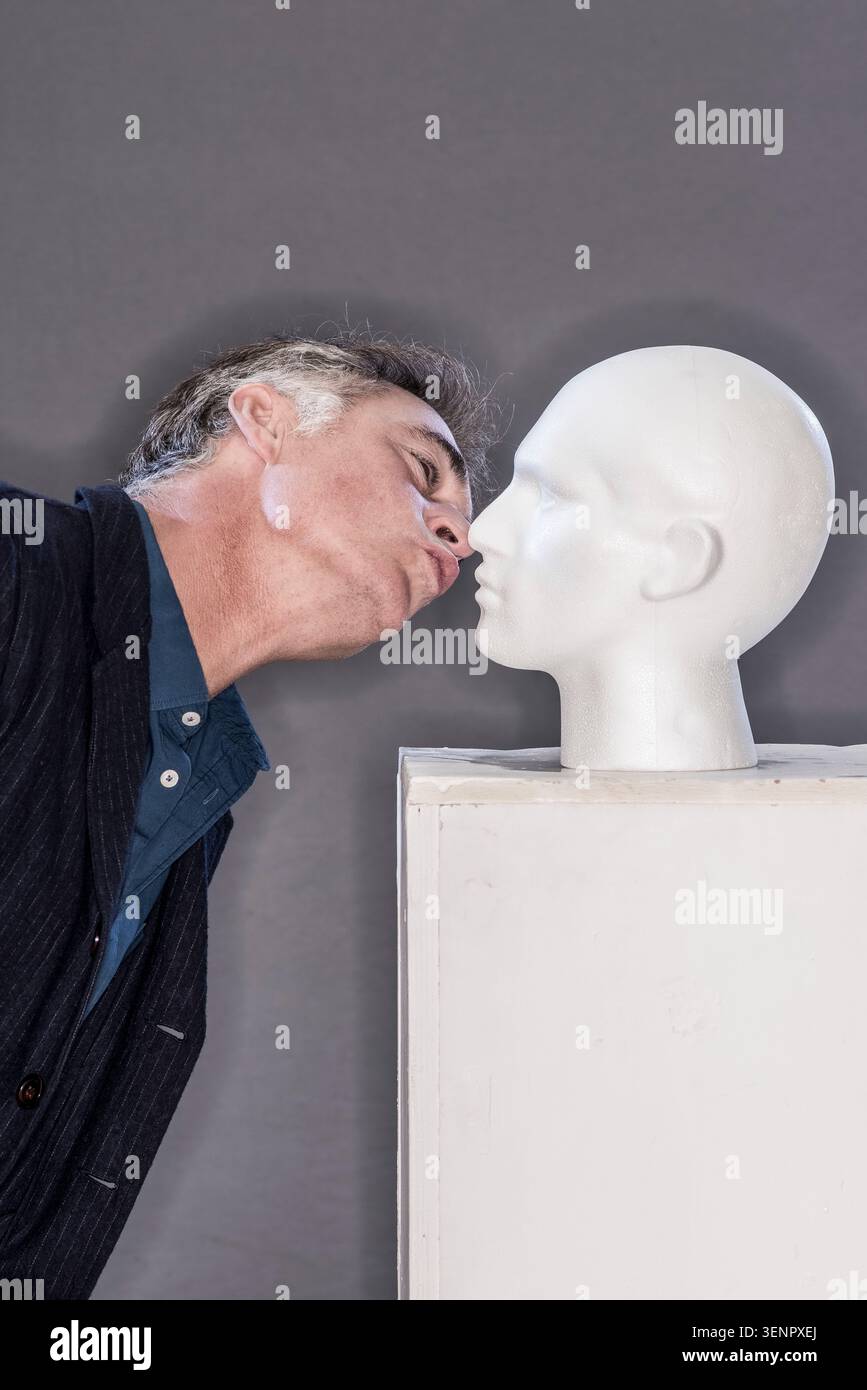 Greg Wise se penche près d'une tête de mannequin blanche dans un studio. Le réglage a un arrière-plan simple, et la lumière est douce. Banque D'Images
