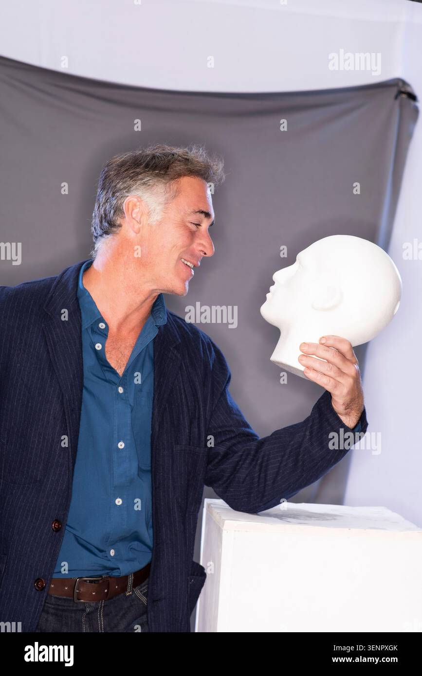 Greg Wise se penche près d'une tête de mannequin blanche dans un studio. Le réglage a un arrière-plan simple, et la lumière est douce. Banque D'Images