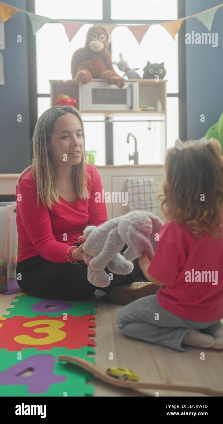 Femme remettant un jouet en peluche à un enfant dans une salle de jeux de bâtiment agenouillée sur un tapis de mousse ; nourrir le collage de jeu. Banque D'Images
