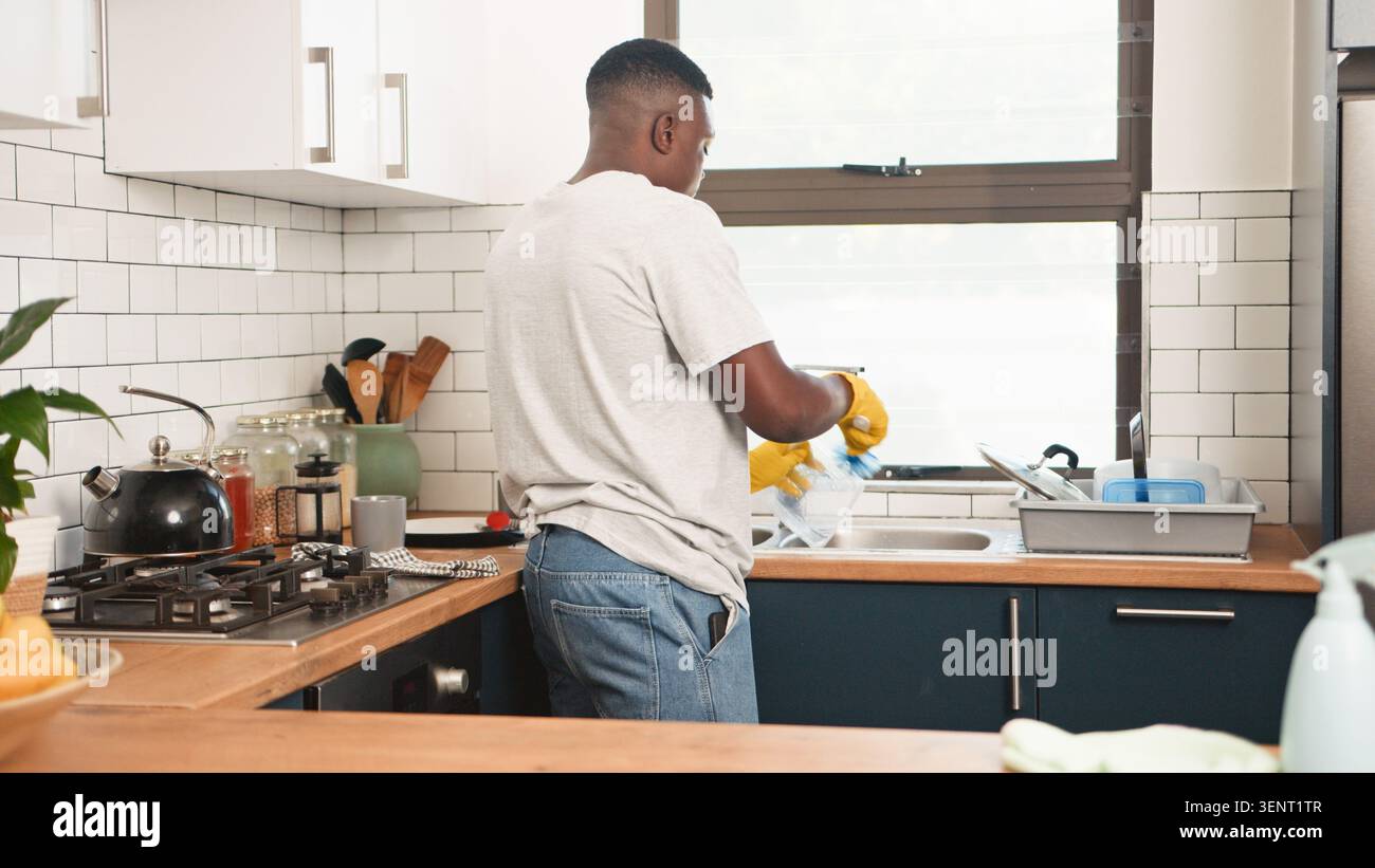 Homme lavant la vaisselle dans la cuisine moderne de la maison portant des gants jaunes et des vêtements décontractés Banque D'Images
