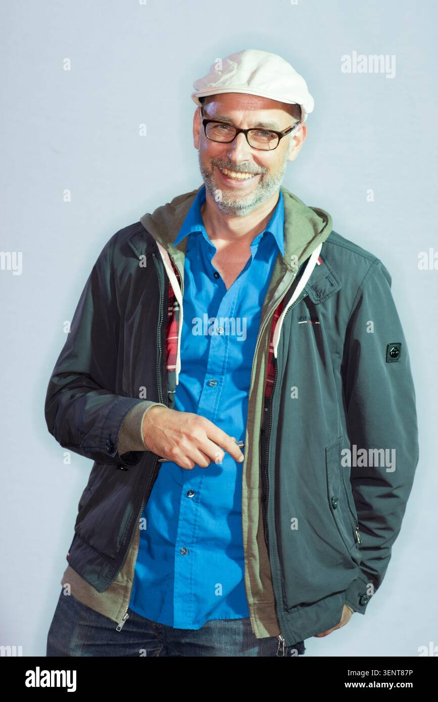Reinhard Kleist pose pour l'appareil photo portant une tenue décontractée. Il sourit habillé d'une chemise bleue, d'une veste et d'un chapeau. L'arrière-plan est clair et Banque D'Images
