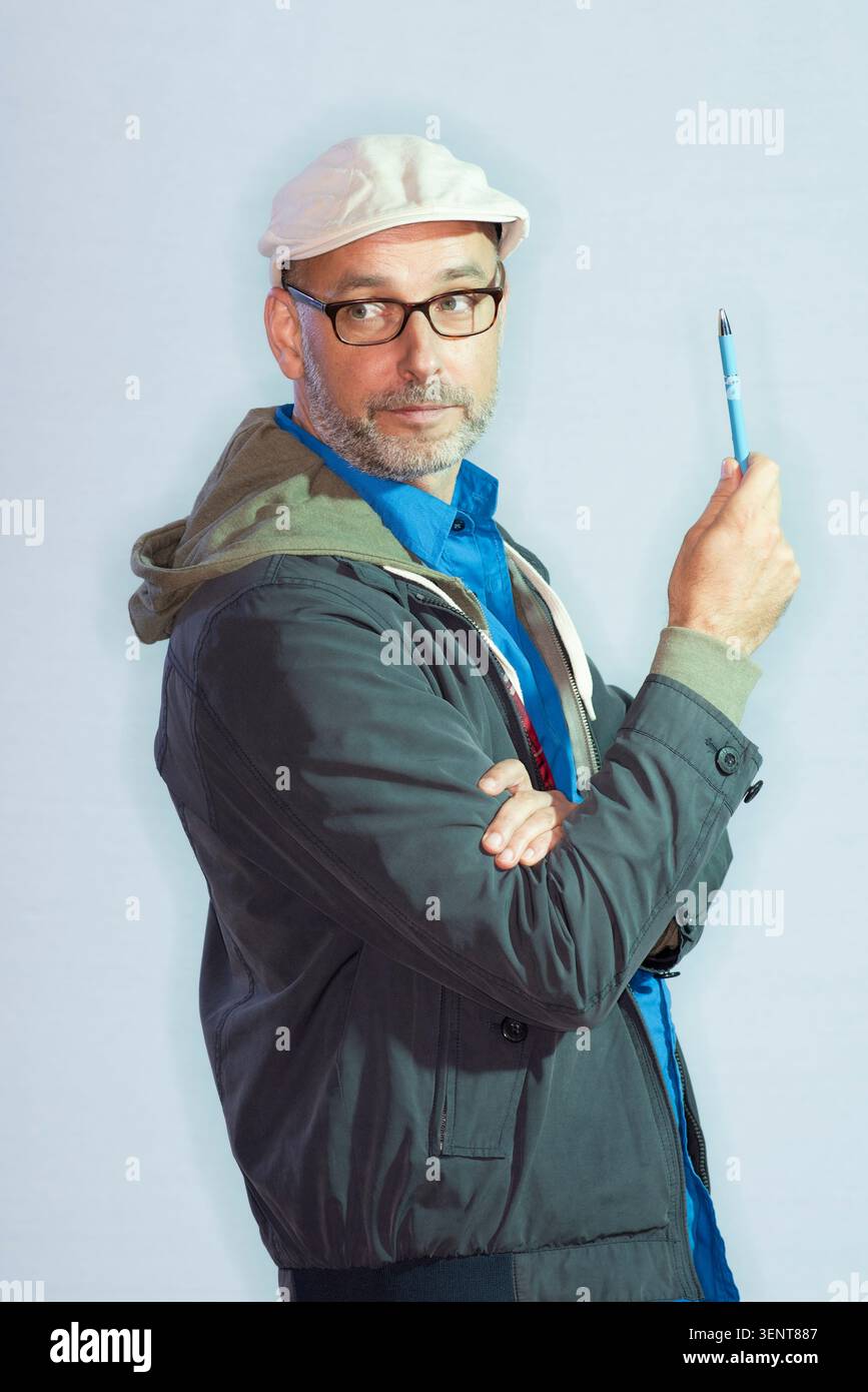 Reinhard Kleist se tient debout dans un environnement de studio, tenant un stylo bleu. Il porte des lunettes et un chapeau, avec un regard sérieux alors qu'il s'engage dans la pensée créative Banque D'Images