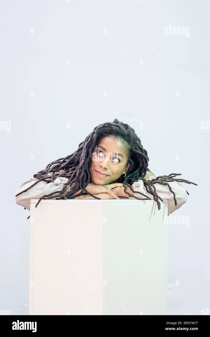 Tomi Adeyemi est engagée dans un moment créatif, reposant ses bras sur un bloc blanc. Elle semble être profonde dans la pensée dans un cadre de studio. Banque D'Images