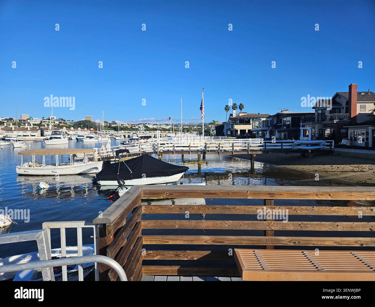 Scène portuaire calme à Balboa Island à Newport Beach, Californie, avec des bateaux amarrés, des maisons en bord de mer, des palmiers, reflets du soleil sur l'eau, et a Banque D'Images