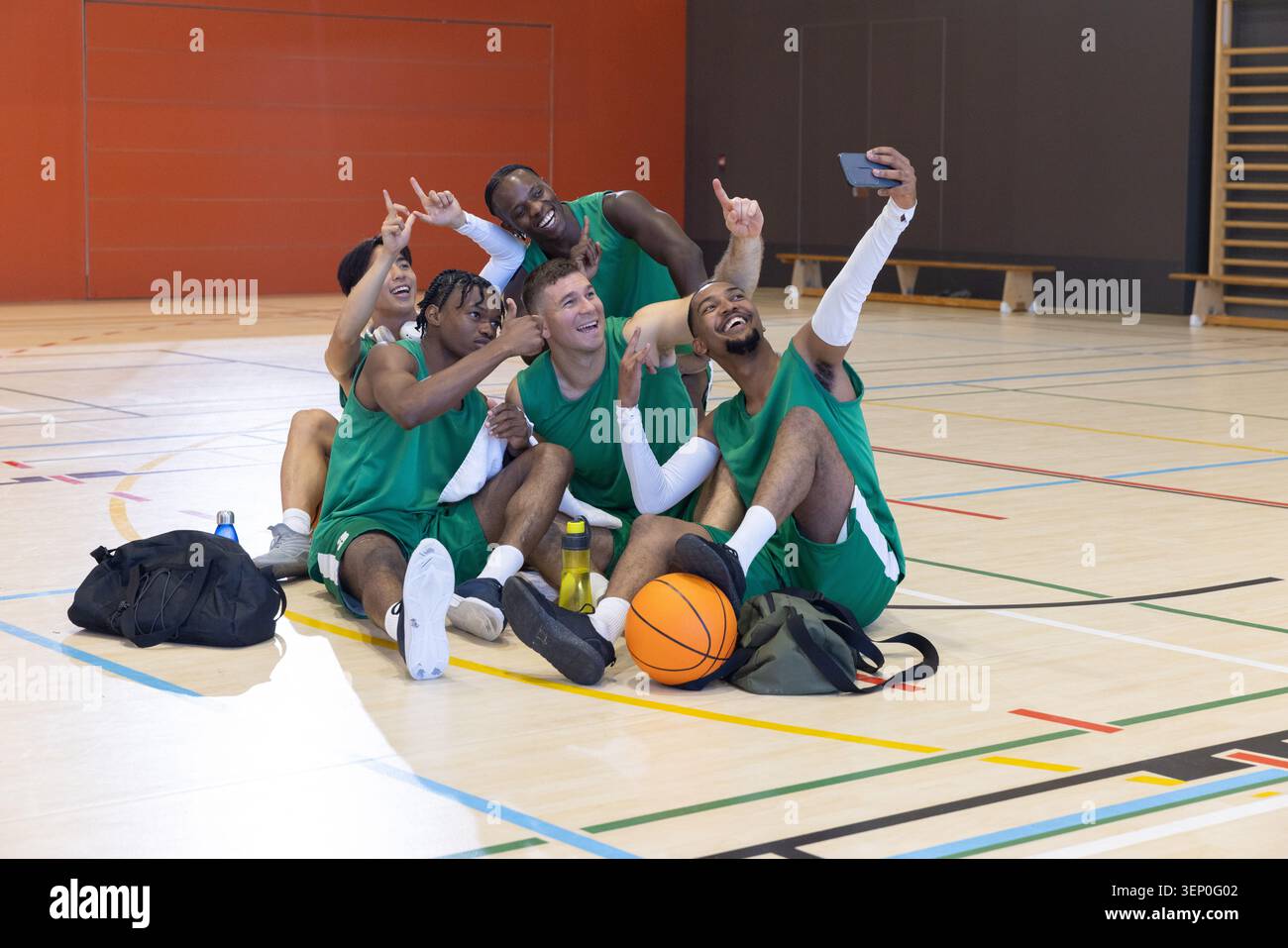 Équipe masculine diversifiée en maillots verts sur le terrain de gymnastique prenant selfie avec téléphone basket-ball et équipement Banque D'Images