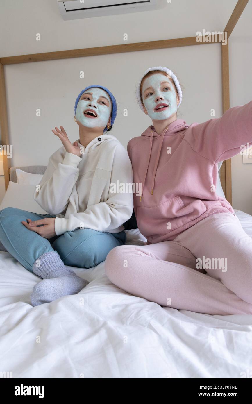 Deux amies assises sur le lit portant des masques faciaux bandeaux en éponge pastel loungewear dans la chambre Banque D'Images