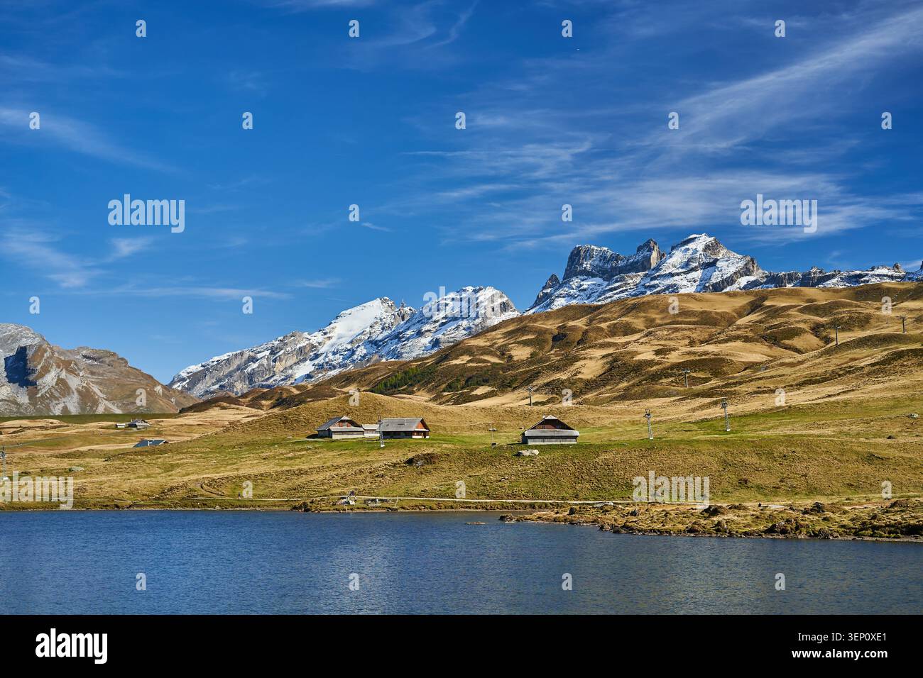 Scène alpine paisible avec un lac calme, des montagnes enneigées, des collines ondulantes et des chalets rustiques dispersés. Banque D'Images