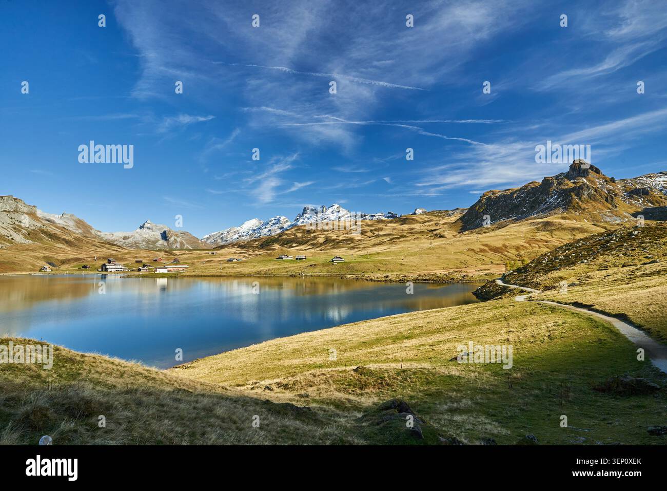 Paysage alpin tranquille avec un lac de montagne réfléchissant, des chalets dispersés et un chemin sinueux. Banque D'Images