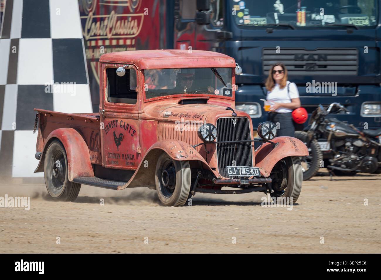 Vue avant d'une camionnette rouge vintage Ford modèle 46 accélérant au début d'une course de dragsters sur la plage, jetant la poussière sur le sable avec un py à carreaux Banque D'Images