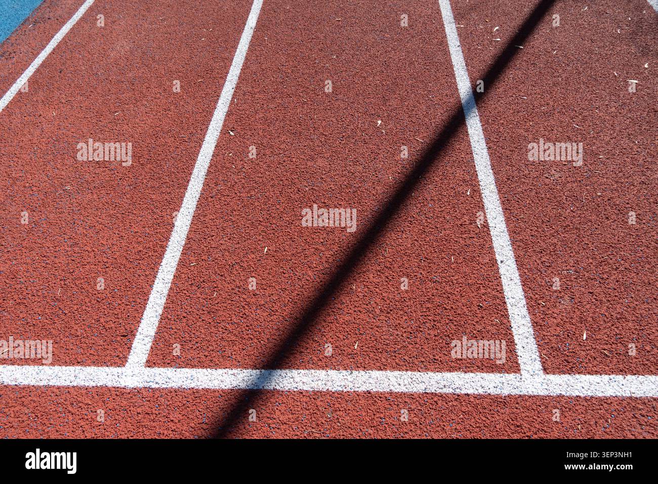 Surface de piste rouge avec lignes blanches distinctes pour la course. Le soleil brille vers le bas, et il y a des feuilles dispersées à la surface. Banque D'Images