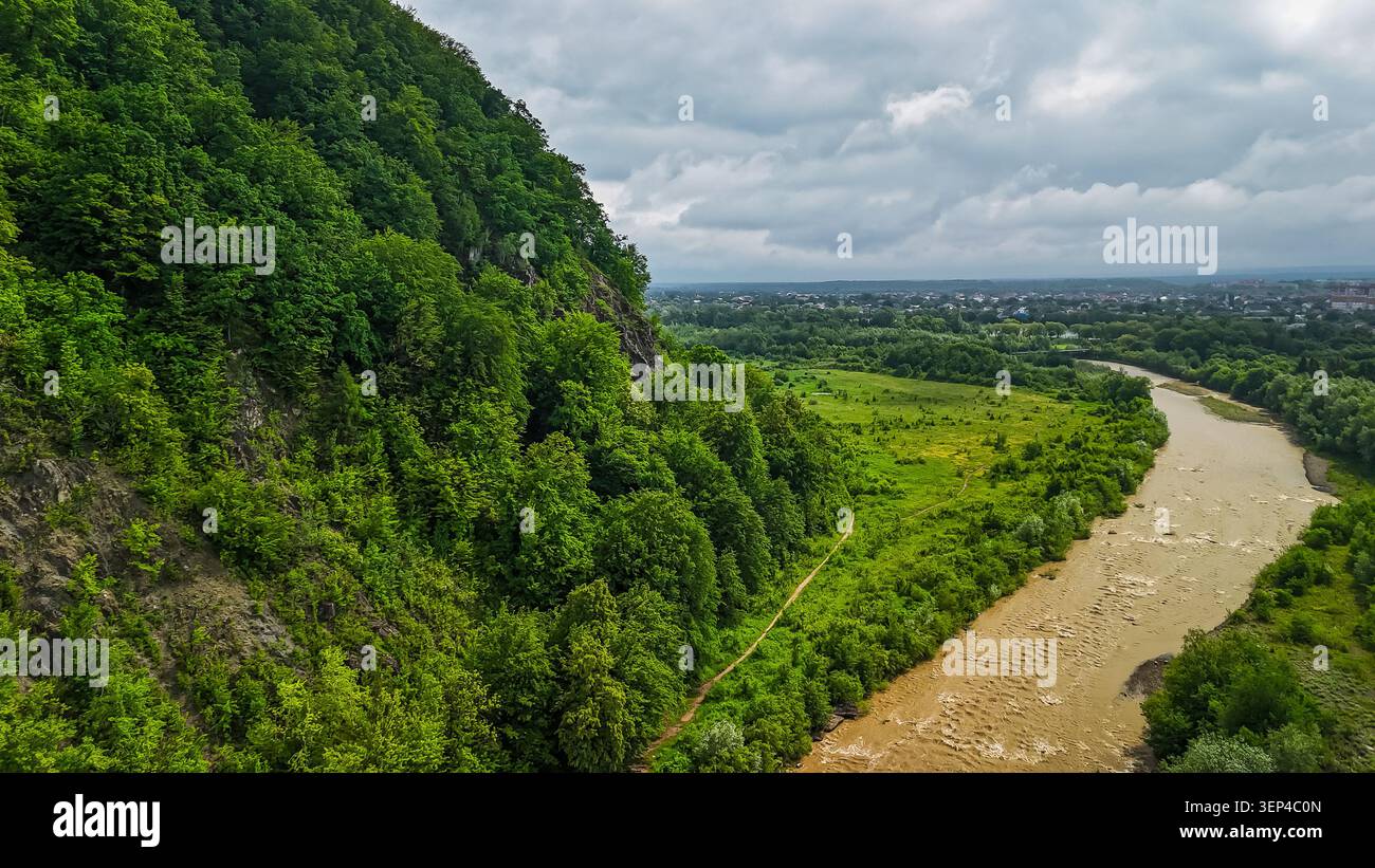 Des falaises abruptes et boisées s'élèvent au-dessus d'une rivière boueuse qui serpente vers une ville lointaine. Drone perspective capture la géologie spectaculaire végétation luxuriante et le c Banque D'Images