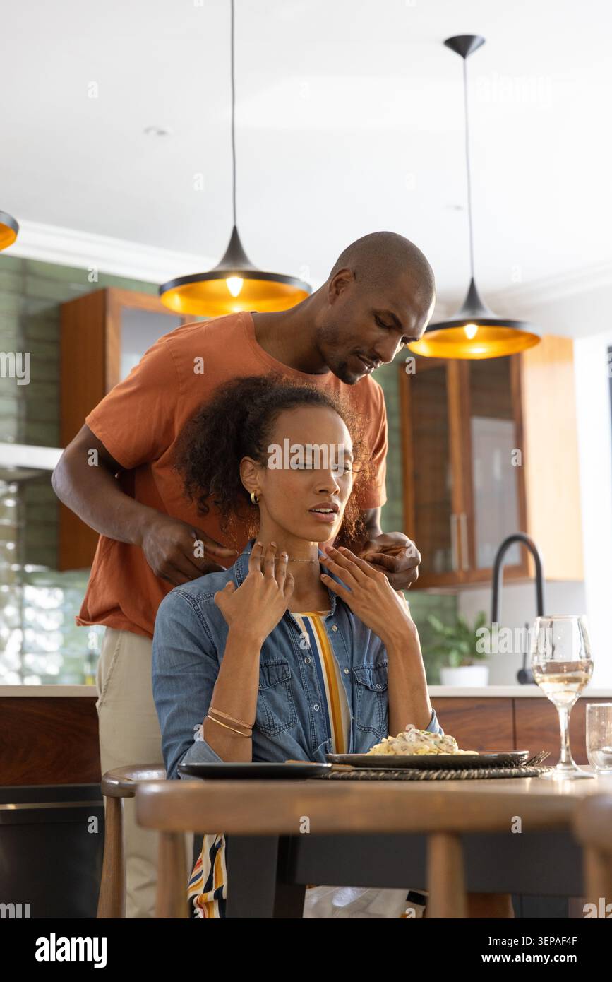 Couple diversifié assis à la table à manger en bois, homme massant le cou de la femme près de l'assiette et verre à vin Banque D'Images