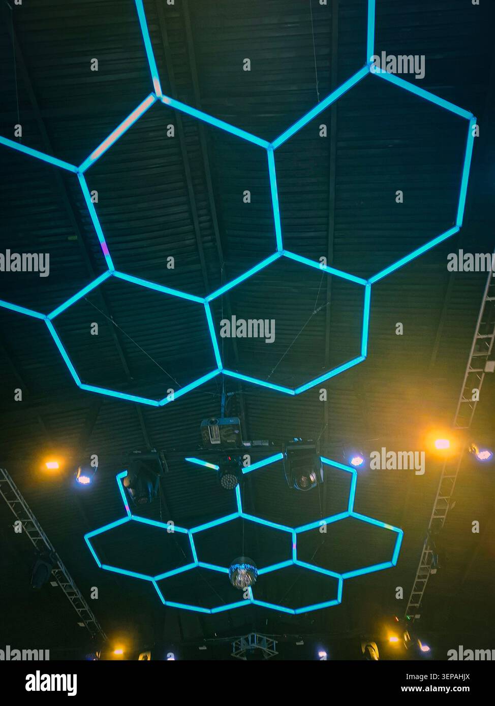 Des lumières LED hexagonales néon bleues pendent d'un plafond industriel sombre avec un équipement d'éclairage de scène présent dans un lieu moderne. Cette configuration est conçue pour Banque D'Images