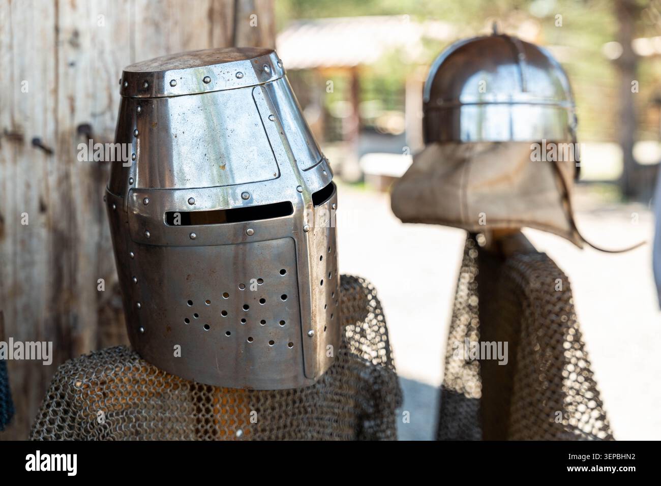 Deux casques de chevalier en métal poli se tiennent à côté de l'armure de mailles de chaîne dans un cadre extérieur avec la lumière du jour douce et un fond rustique flou, suggérant un Banque D'Images
