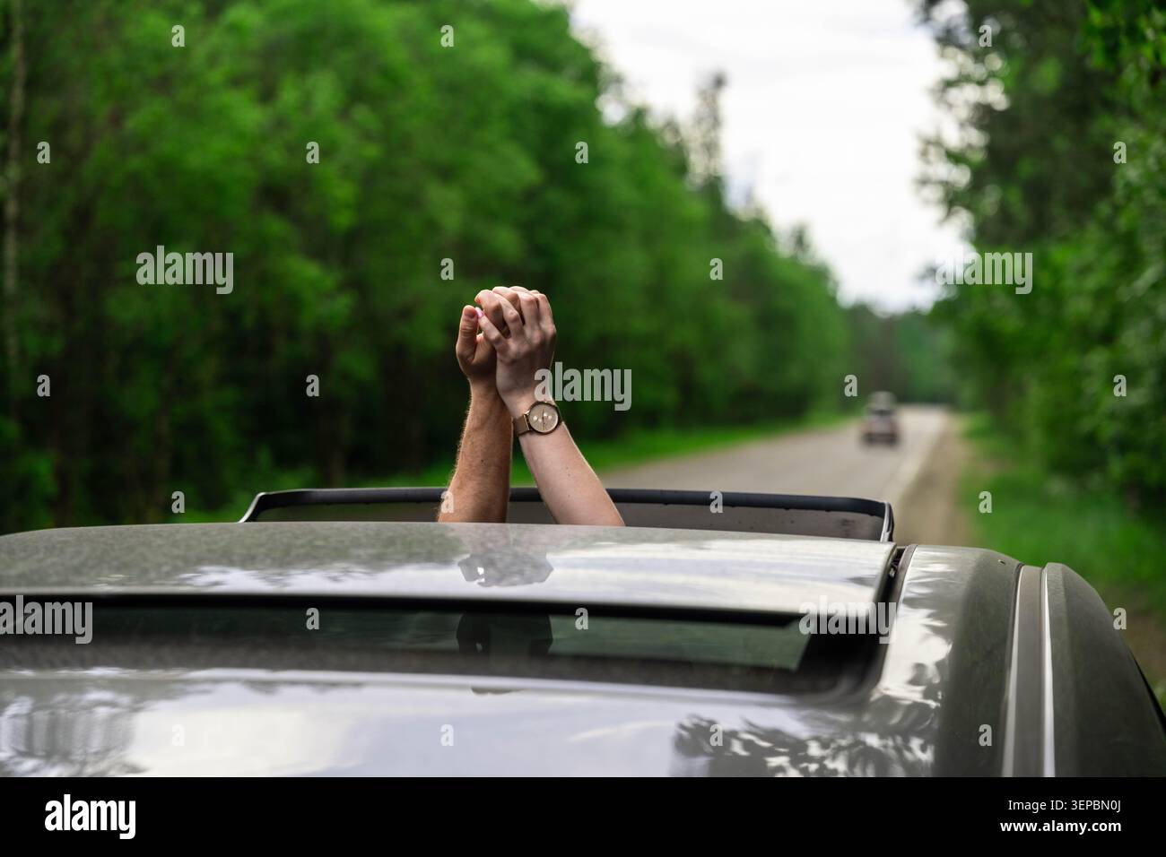 Paire de mains serrées s'étend à travers le toit ouvrant ouvert de la voiture en mouvement sur la route bordée d'arbres, avec un bois vert flou et un ciel couvert en arrière-plan Banque D'Images