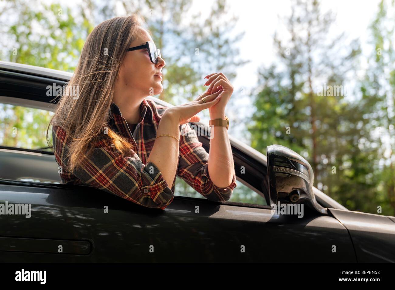 Jeune femme portant des lunettes de soleil et une chemise à carreaux se penche par la fenêtre de la voiture avec ses mains serrées, entourée d'arbres verts et d'une lumière naturelle éclatante Banque D'Images
