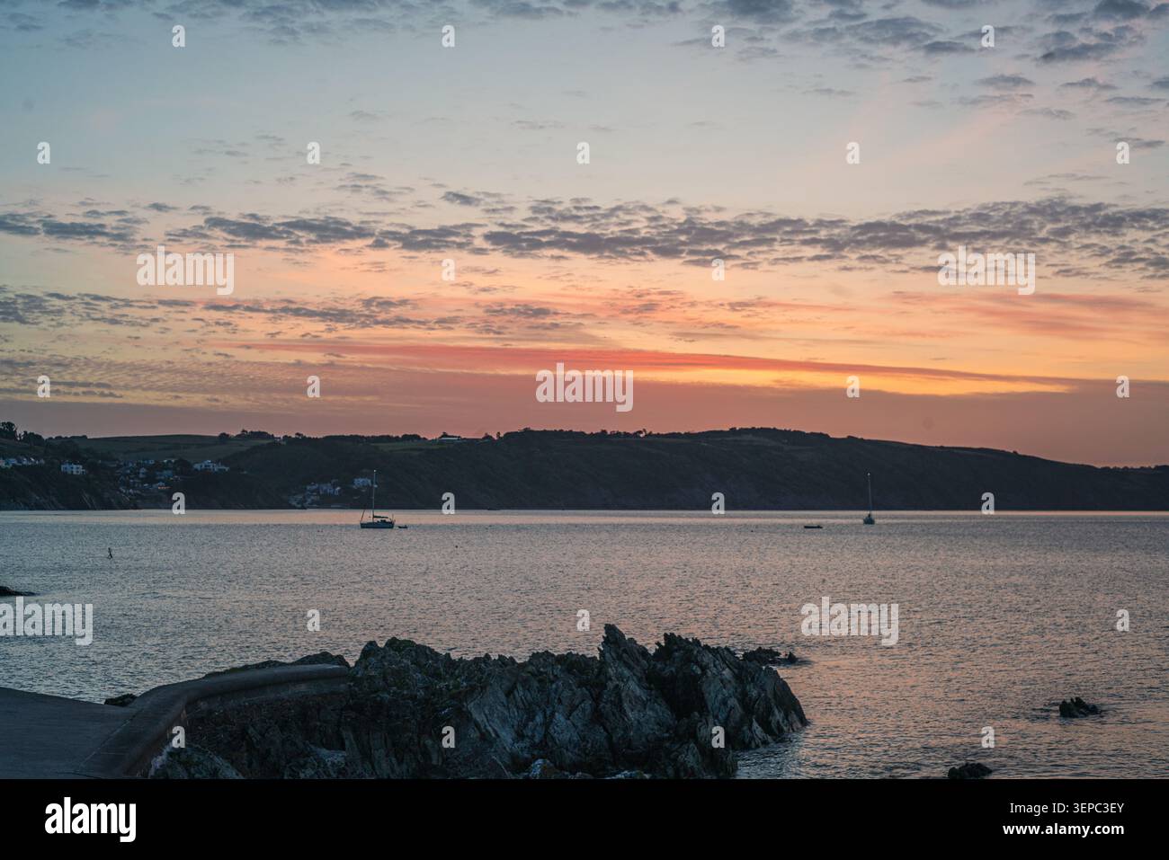Ciel éclatant du matin aux couleurs pastel douces au-dessus de l'eau calme, avec un voilier ancré près d'une structure rocheuse sombre au premier plan à Looe Banque D'Images