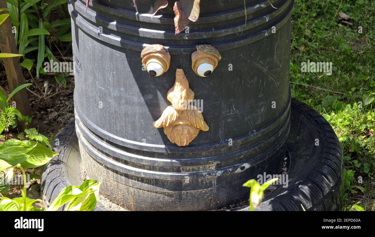 Une jardinière en plastique noir texturé en forme de tonneau est décorée d'un visage comique fait d'un nez en bois, d'une moustache et de grands yeux googly assis Banque D'Images