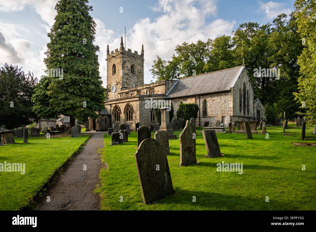 Vue horizontale large de l'église Saint-Laurent montrant la grande tour de pierre, les pierres tombales du cimetière et un sentier pédestre sous un ciel nuageux d'été à Eyam Banque D'Images