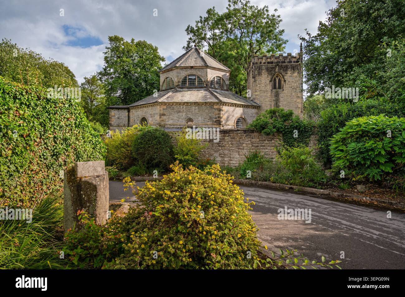 Vue panoramique horizontale sur l'église historique St Martin encadrée par un poteau de pierre et des buissons verdoyants de jardin à Stoney Middleton Banque D'Images