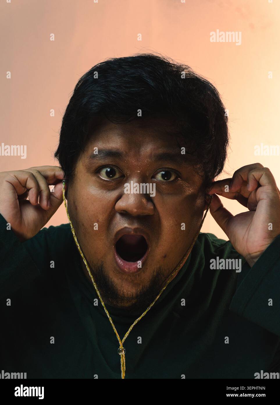 Un homme indonésien portant une chemise vert foncé insère des écouteurs filaires dans ses oreilles tout en regardant l'appareil photo avec une expression intense choquée contre A. Banque D'Images