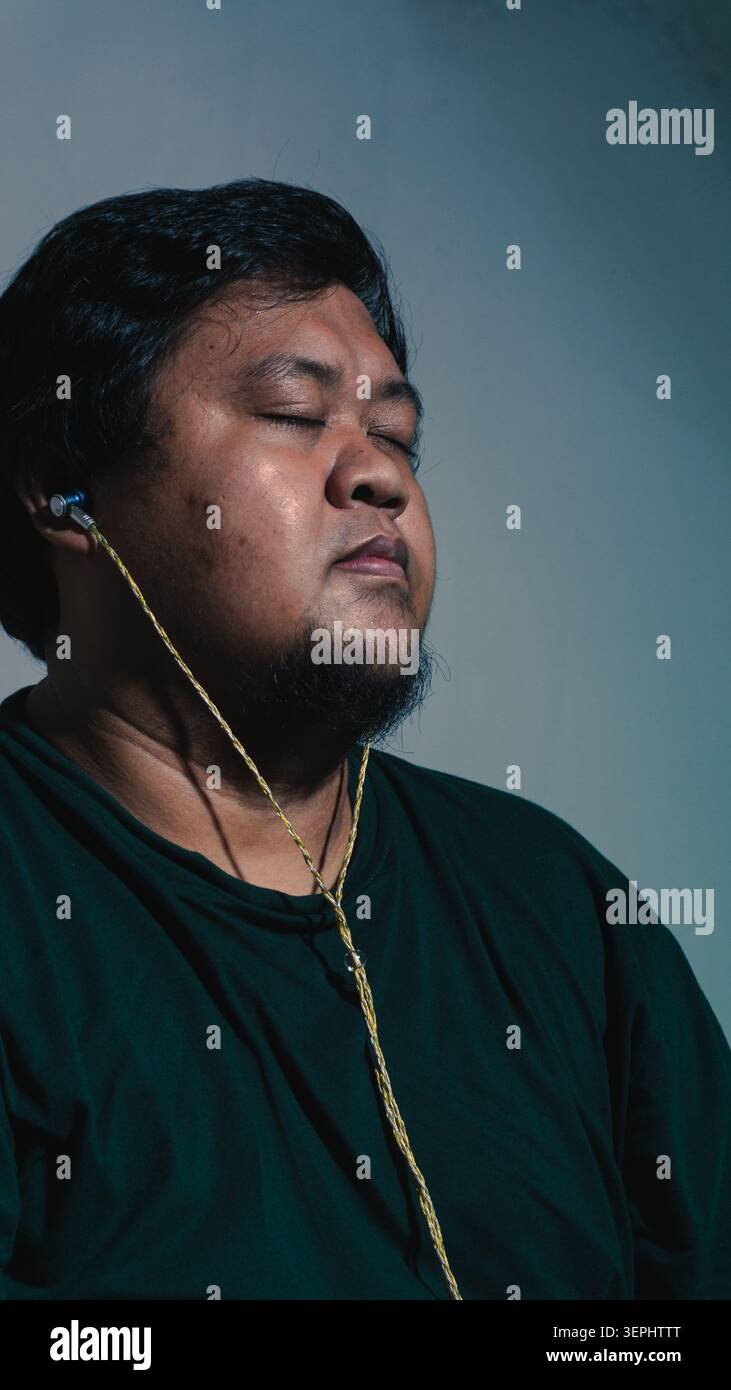 Un homme indonésien portant une chemise vert foncé garde les yeux paisiblement fermés tout en écoutant de la musique à travers des écouteurs filaires contre un BA gris frais Banque D'Images