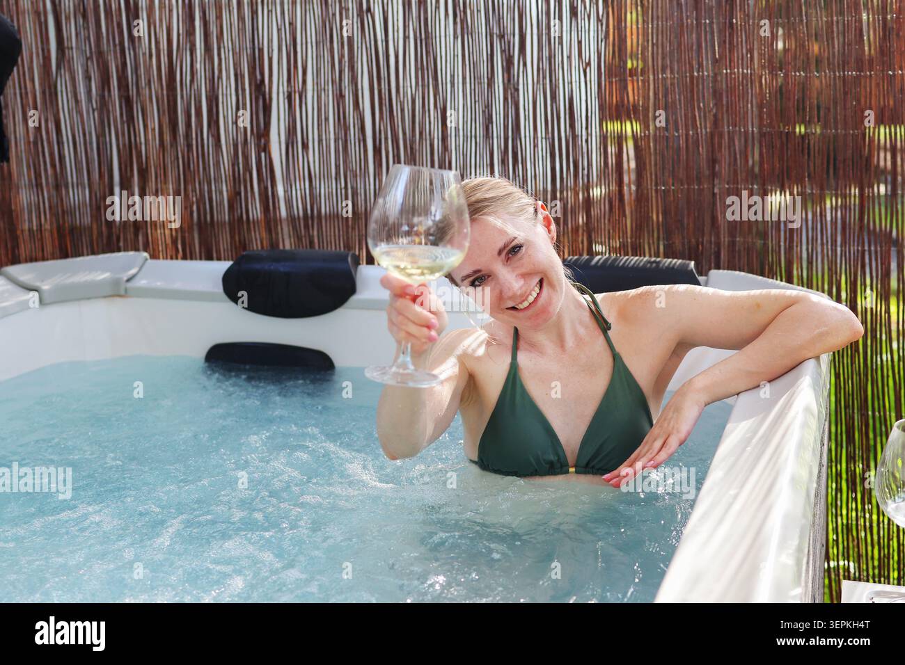 Jeune femme célébrant le temps de vacances dans un bain à remous moderne avec de l'eau pétillante et un verre de vin blanc. Banque D'Images