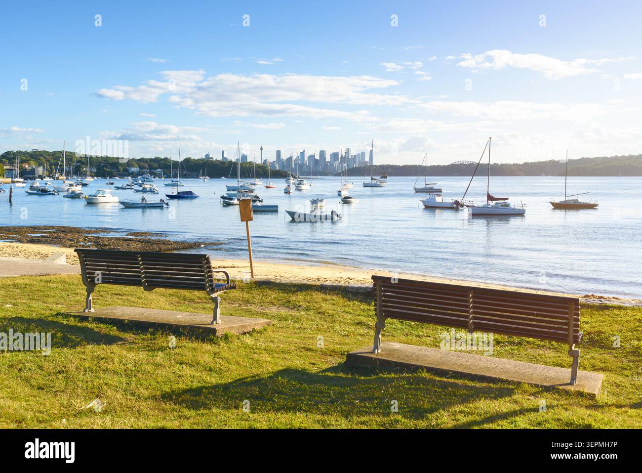 Bancs en bois vides sur l'herbe face à une baie parsemée de bateaux ancrés par une journée ensoleillée d'automne. L'horizon de Sydney est visible en arrière-plan. Banque D'Images