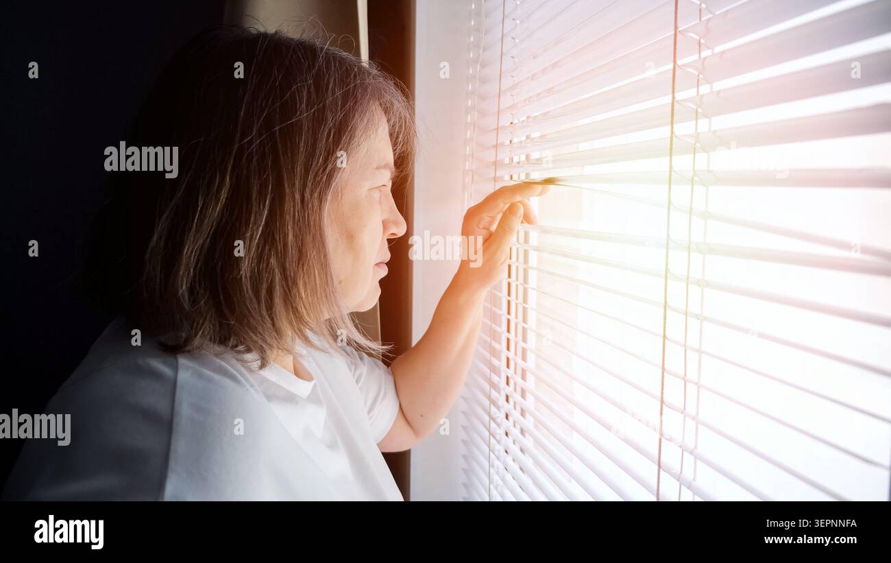 Femme âgée debout près d'une fenêtre, regardant à travers des stores horizontaux avec ses doigts, réfléchissant sur les concepts de solitude, d'isolement et d'espoir Banque D'Images