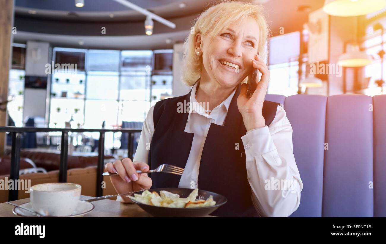 Femme d'affaires âgée caucasienne souriant au téléphone tout en mangeant de la salade et en buvant du café au Sunlit Cafe Booth, tenant stylo et notes de note, détendue Banque D'Images