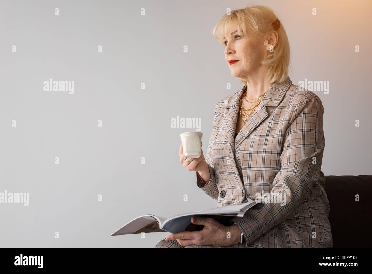 Femme blonde mature portant un blazer à carreaux, tenant un magazine et une tasse, regardant loin contemplativement, perdu dans la pensée en attendant ou en prenant un Banque D'Images