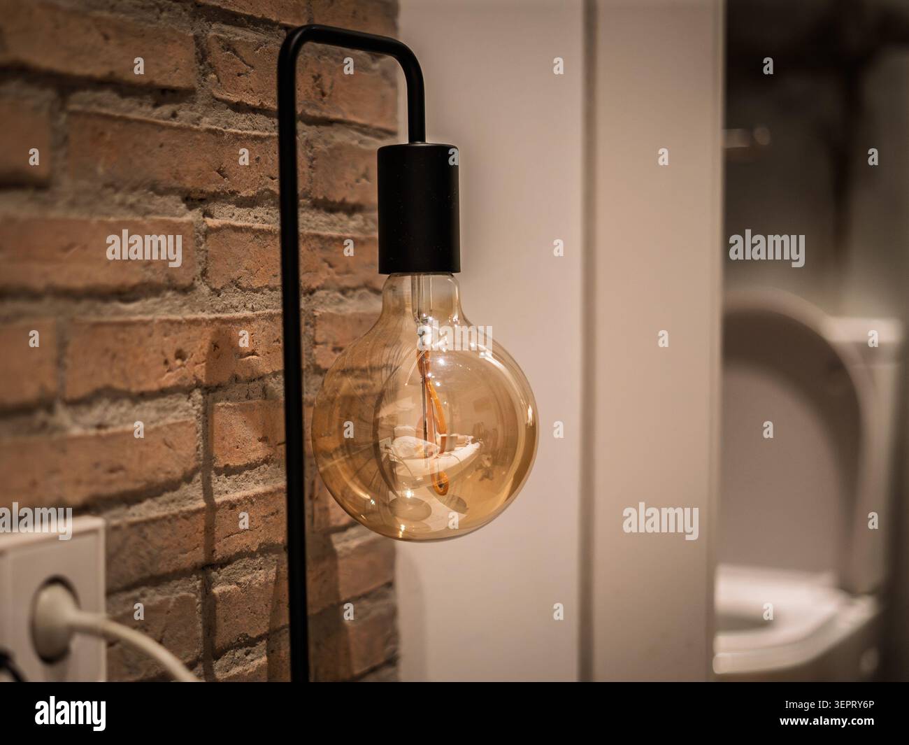Une ampoule incandescente vintage est suspendue à un appareil mural noir à côté d'un mur de briques apparentes, avec un globe en verre ambré, filament visible, lueur chaude Banque D'Images