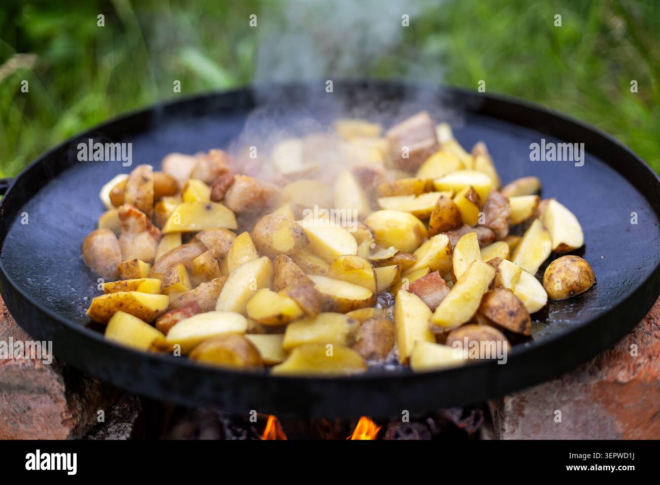 Cuisson des pommes de terre et du porc dans une poêle fumée extérieure. Des quartiers de pommes de terre dorées et des morceaux de porc grésillent dans une poêle en fonte noire au-dessus d'une flamme nue, avec Banque D'Images