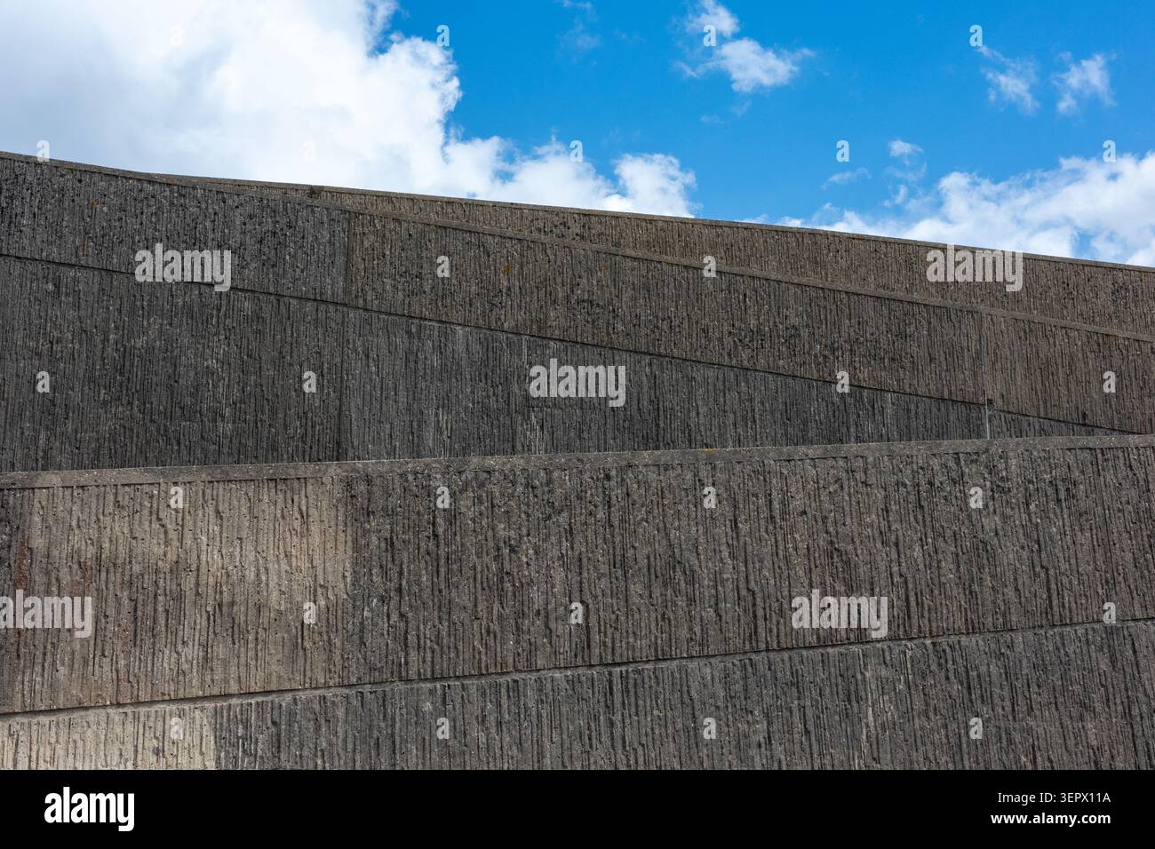 Un mur de béton en pente est vu sur fond de ciel bleu et de nuages blancs. La texture et les lignes de la structure créent un design urbain clair. Le SC Banque D'Images
