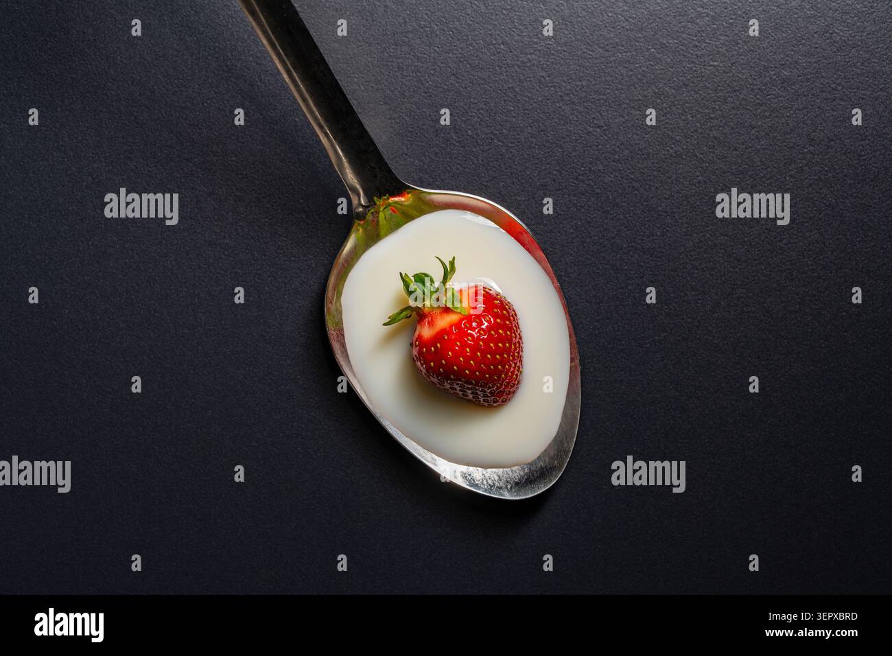 Fraise fraîche avec yaourt blanc dans une cuillère sur fond noir, gros plan. Vue de dessus d'une seule fraise rouge mûre à l'intérieur d'une cuillère en métal remplie de Banque D'Images