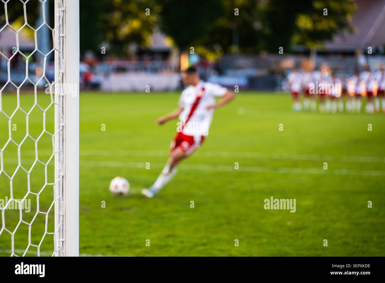 Un joueur de football dans un uniforme blanc frappant un tir de pénalité vers le but pendant un match de football, vu à travers le filet. Banque D'Images