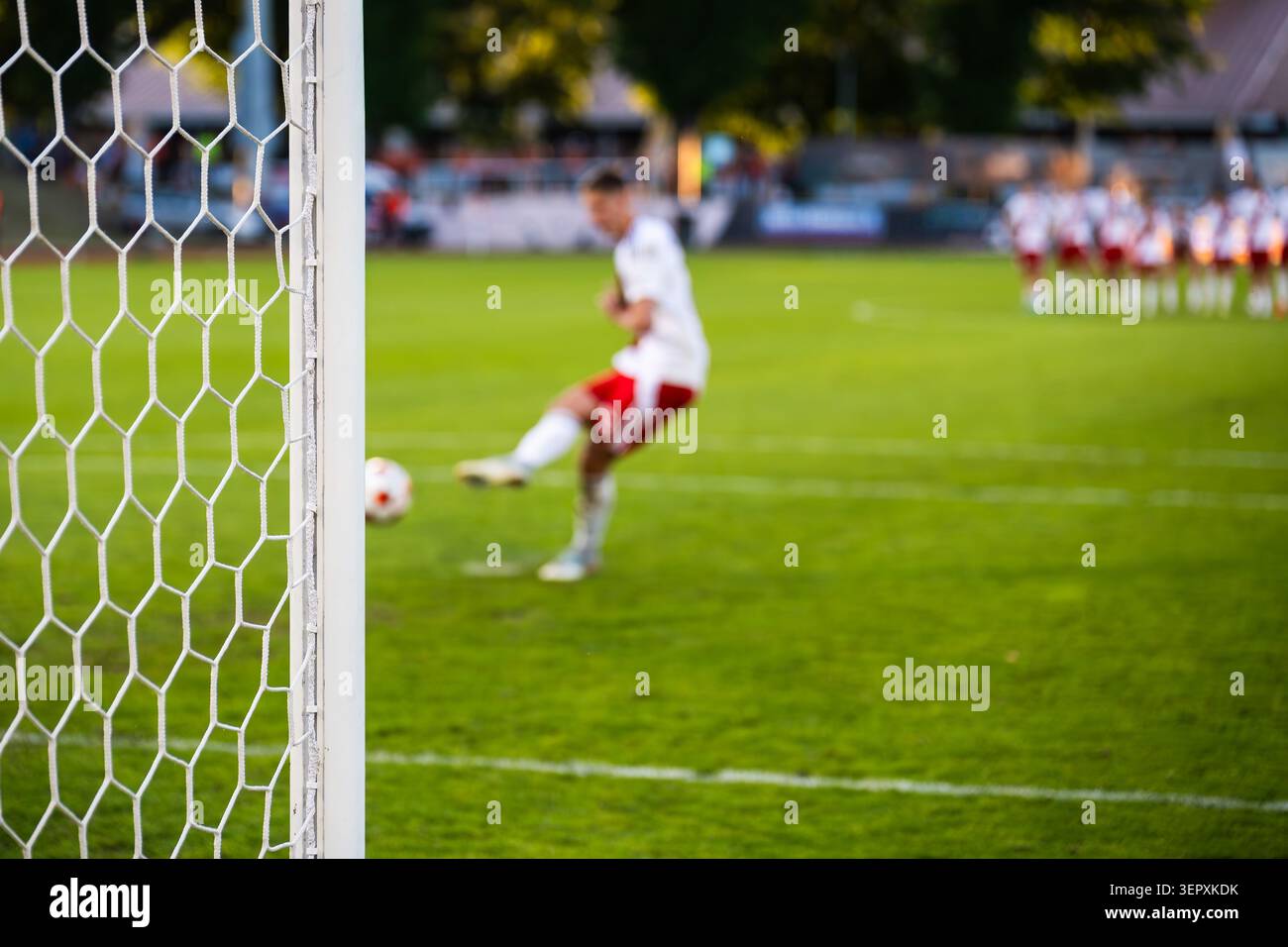 Un joueur de football dans un uniforme blanc frappant un tir de pénalité vers le but pendant un match de football, vu à travers le filet. Banque D'Images