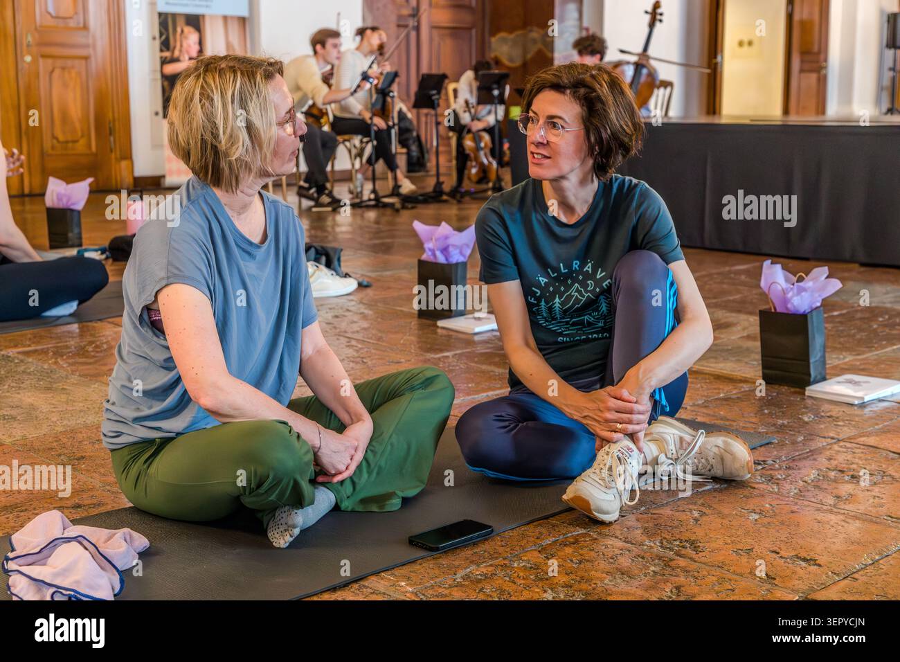 Andrea Stockhammer (à droite) et Angela Berg (à gauche) participent à l’événement « Pop Up Yoga – Mozart Edition » au DomQuartier de Salzbourg. Le réalisateur est assis sur un tapis de yoga parlant au journaliste ; un ensemble de chambre joue en direct en arrière-plan. L’événement s’inscrivait dans le cadre du programme Mozart 270 pour marquer le 270e anniversaire de la naissance du compositeur (1756-2026). Domplatz, Salzbourg, Autriche Banque D'Images