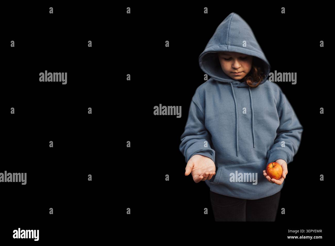Enfant vulnérable portant un sweat à capuche tenant une petite pomme et tendant une main vide dans l'obscurité totale symbolisant la pauvreté Banque D'Images