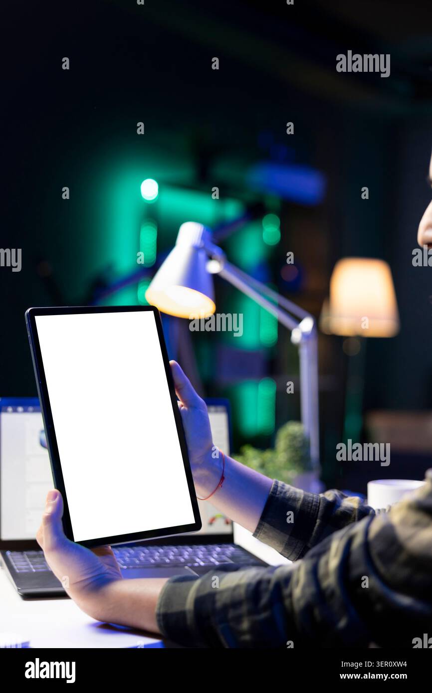 Personne tenant verticalement la tablette avec écran blanc devant l'ordinateur portable. Freelance masculine travaillant à la maison, axée sur l'appareil numérique affichant un modèle vierge de maquette de clé chroma. Banque D'Images