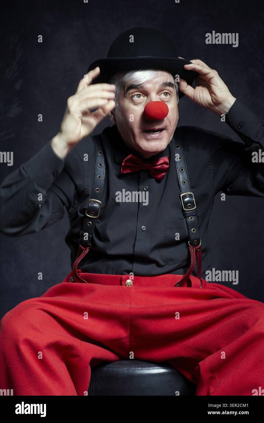 Portrait d'un clown sur fond sombre. Un homme âgé avec un nez de clown rouge et un chapeau melon sur la tête. Vêtu d'un pantalon rouge et d'une chemise noire avec suspension Banque D'Images