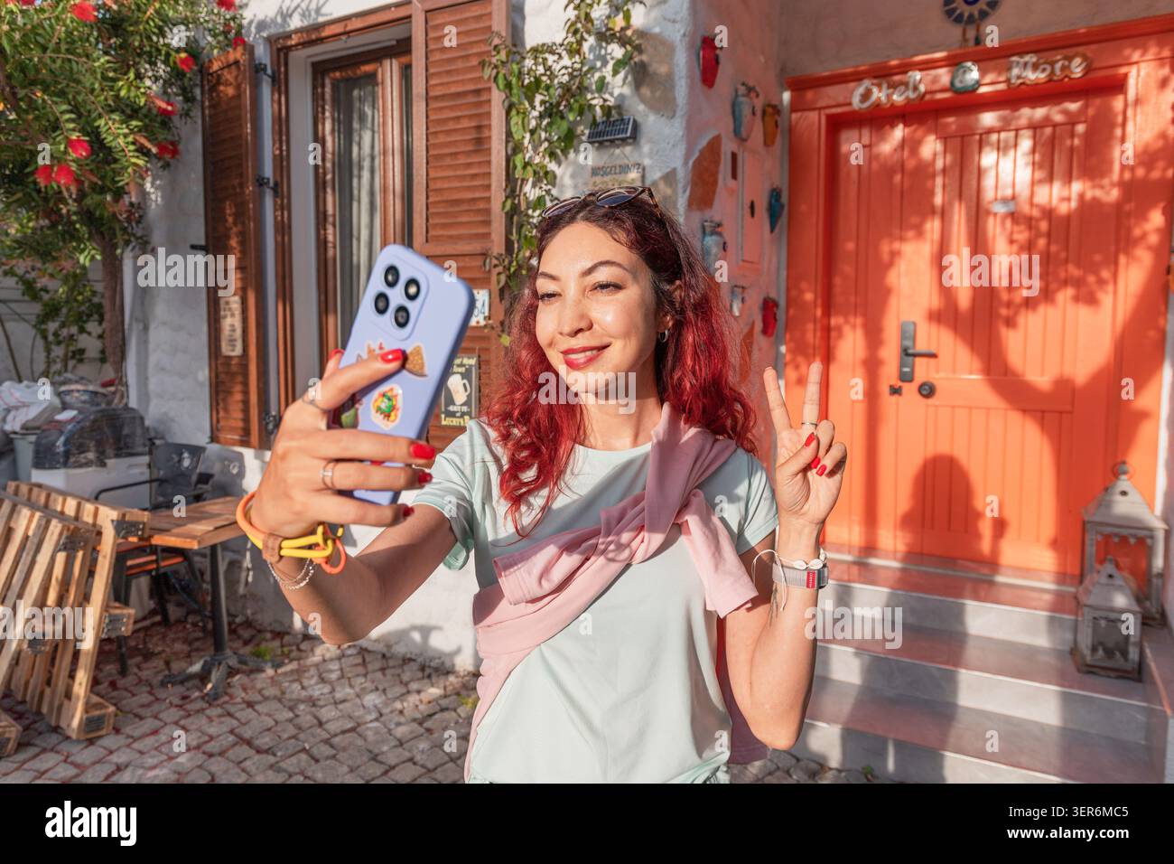 13 mai 2026, Sigacik, Turkiye : femme aux cheveux roux souriant, tenant un téléphone pour un selfie avec un signe de paix devant une porte orange, voyageant Banque D'Images