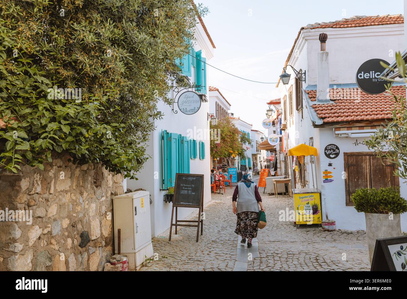 13 mai 2026, Sigacik, Turkiye : femme marchant le long d'une ruelle pavée traditionnelle bordée de maisons blanches et de volets turquoise Banque D'Images